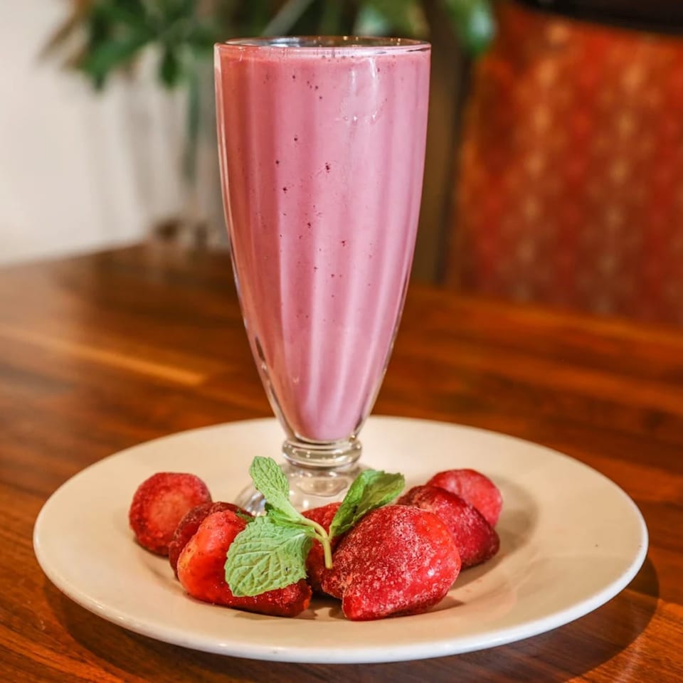 A tall glass of pink vegan strawberry milkshake with frozen strawberries and mint on a plate.