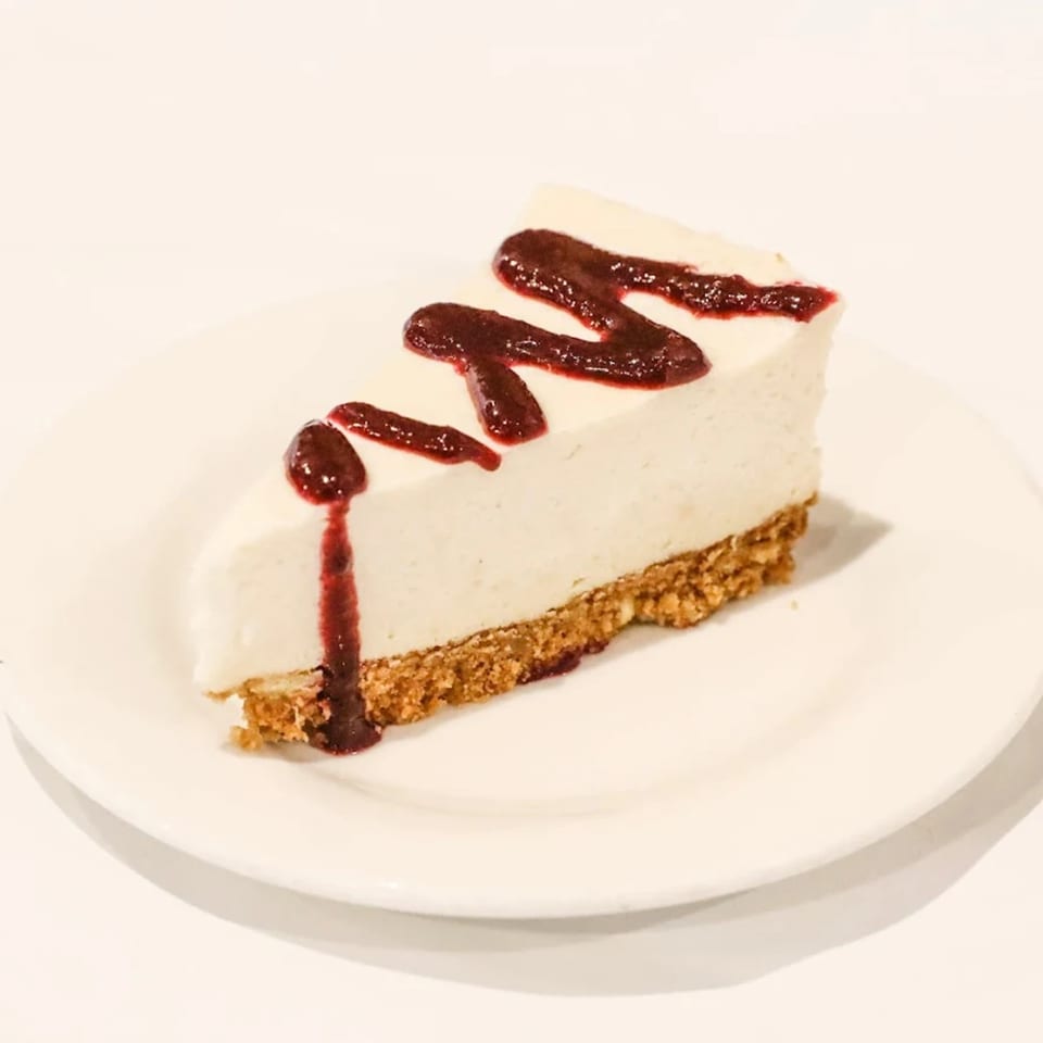 A slice of Blossom Vegan cheesecake with a graham cracker crust and berry drizzle on a white plate.