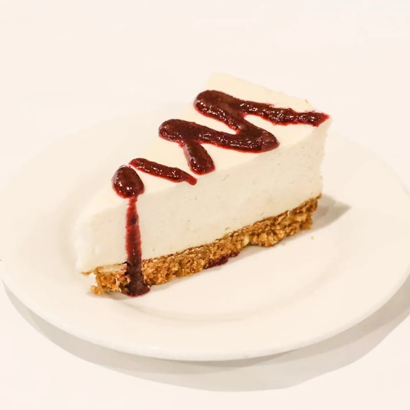 A slice of Blossom Vegan cheesecake with a graham cracker crust and berry drizzle on a white plate.