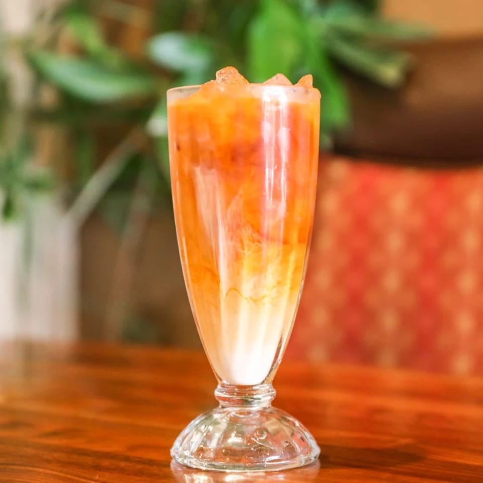 A tall glass of iced vegan Thai tea with a creamy white base and orange tea swirling above.